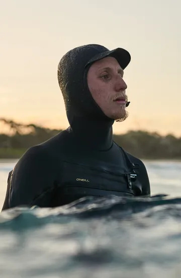 Capucha de neopreno negra para deportes acuáticos, usada por un surfista parcialmente sumergido al atardecer.