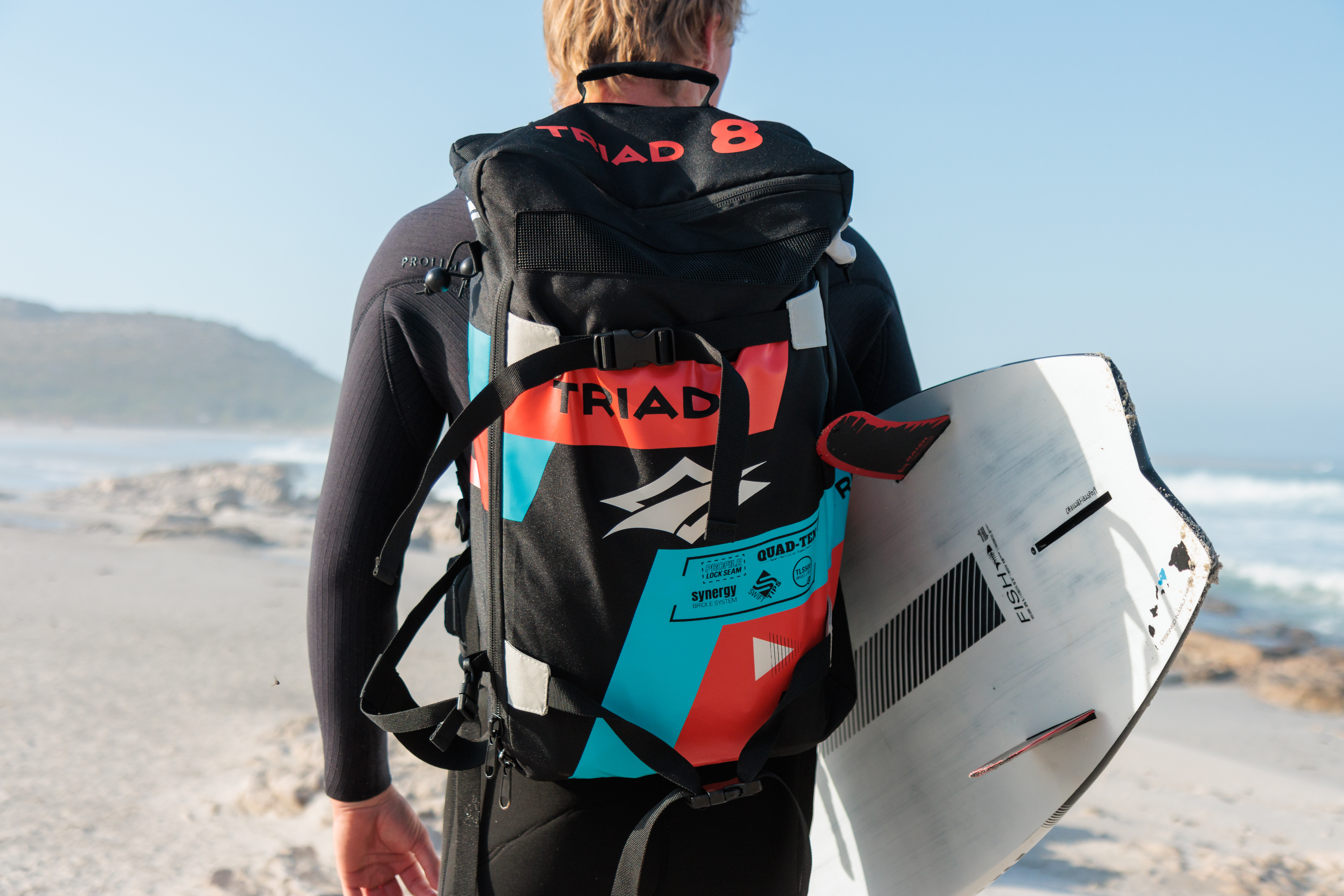 Kitesurfista con mochila técnica y tabla de kitesurf listo para entrar al agua en una playa ventosa