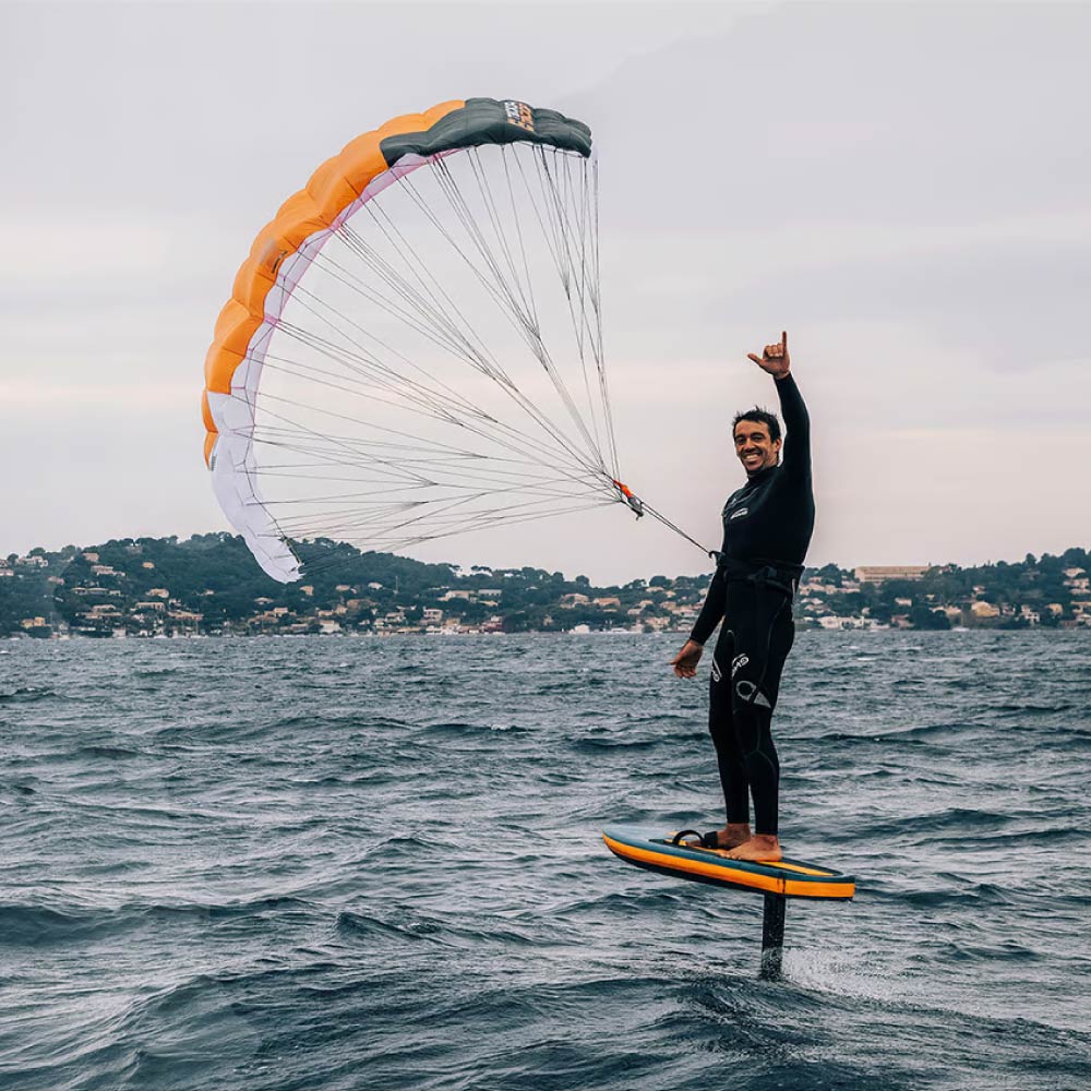 Sportivo in mare che pratica il parawing con tavola hydrofoil e vela arancione e bianca, davanti alla costa