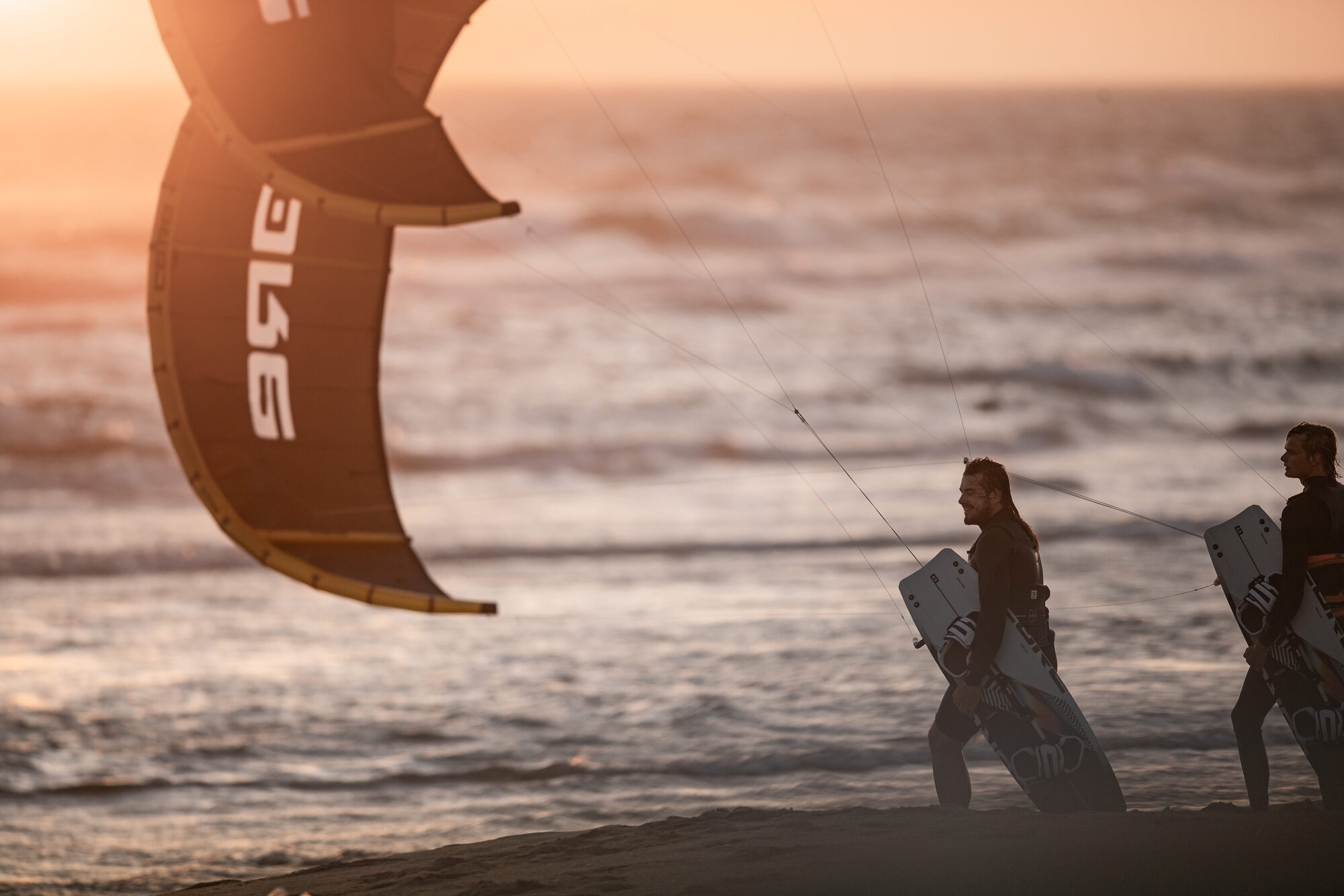 Dos kitesurfistas principiantes caminando por la playa al atardecer con tabla y cometa listos para entrenar
