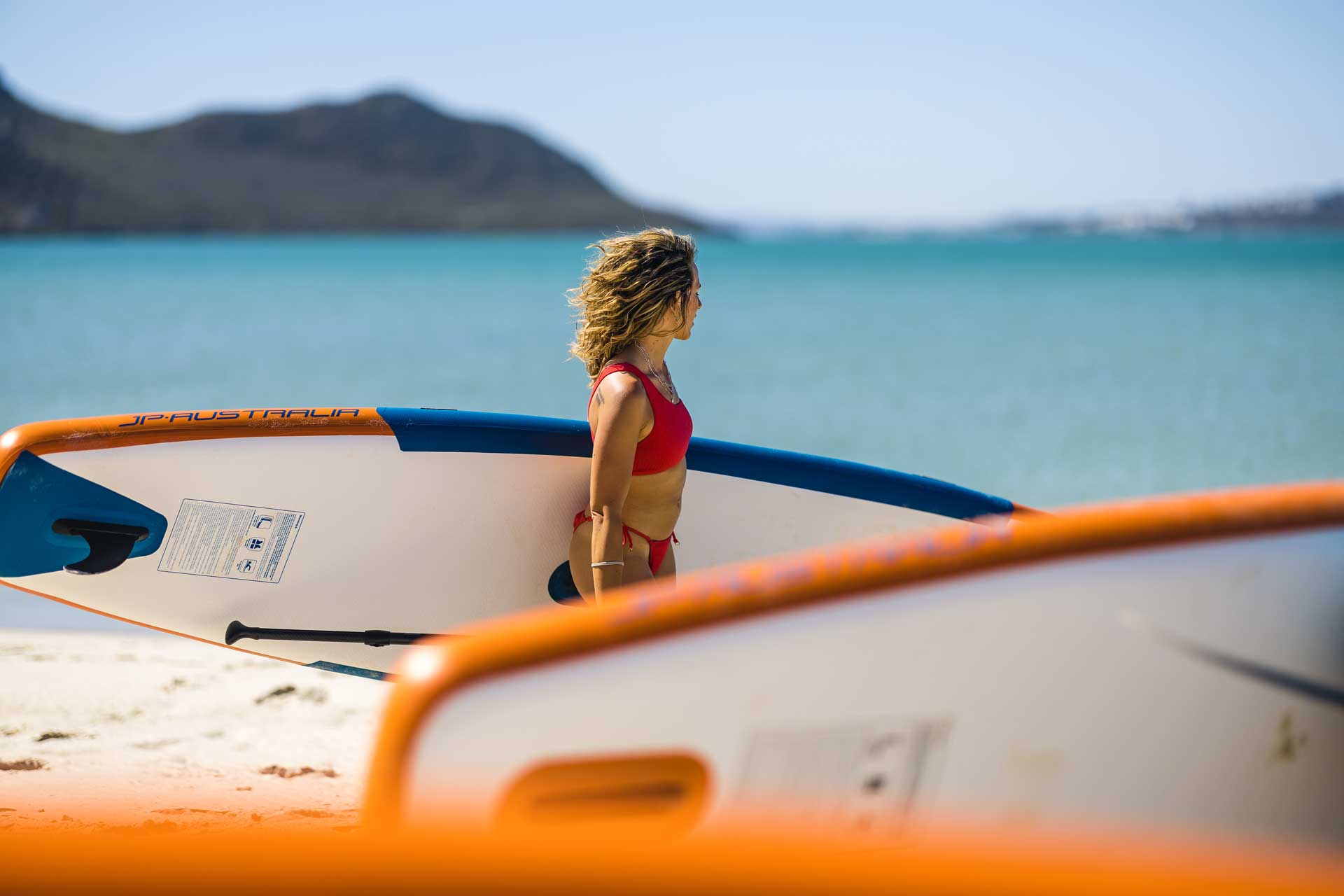 Woman in a red bikini carrying a JP Australia SUP board on the beach in front of turquoise sea