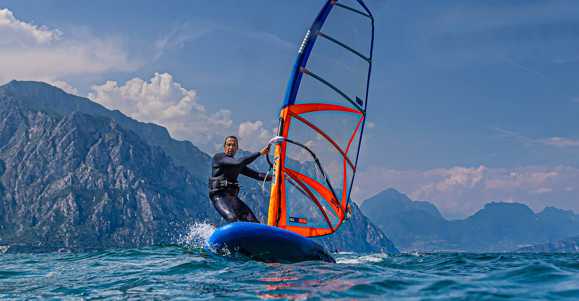 Man windsurfing on a convertible SUP board with an orange sail on a lake, surrounded by mountains.