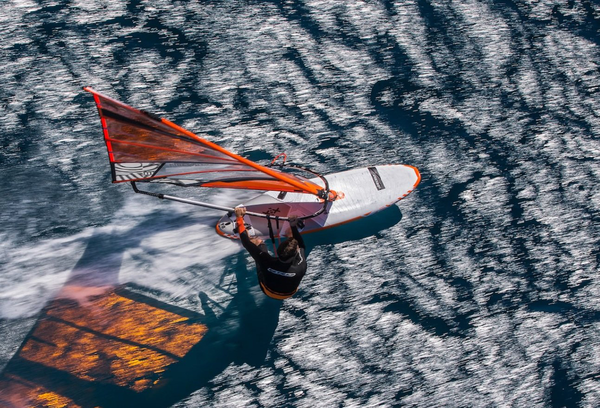Windsurfer con vela arancione e bianca visto dall'alto mentre naviga su acqua increspata.